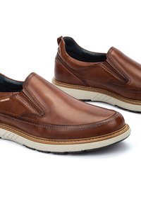 Zapatos marrones de cuero tipo slip-on con un acabado suave, detalles de costura y una suela de goma clara en contraste. Cuentan con paneles elásticos a los lados para poner y quitar fácilmente.