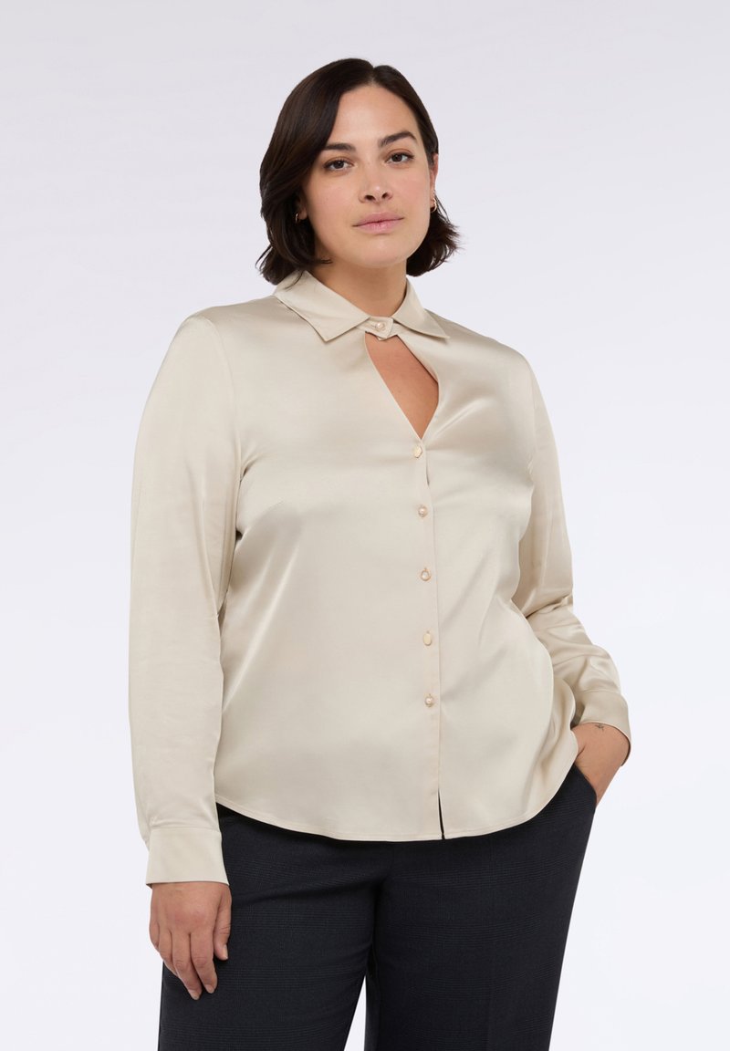 Camicia in satin di colore crema, a maniche lunghe, con colletto, un'apertura a forma di chiave sul petto e bottoni perlacei lungo il davanti. Tessuto liscio e design aderente.