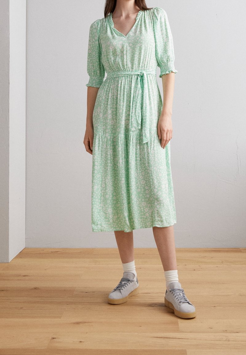Femme portant une robe midi florale vert clair avec taille nouée, manches mi-longues, chaussettes blanches et baskets grises, sur un sol en bois et un mur uni.