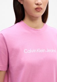Gaiši rozā kokvilnas T-krekls ar apaļu kakla izgriezumu un īsām piedurknēm. Priekšpusē ir drukāts teksts "Calvin Klein Jeans" baltā krāsā.