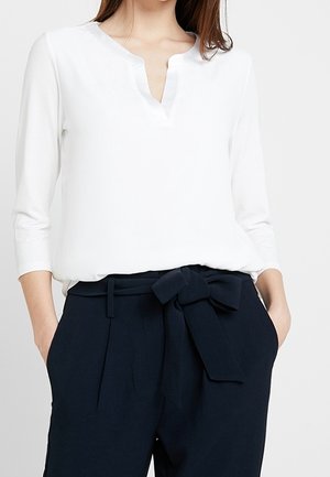 Femme portant un chemisier blanc à manches longues avec un col en V, rentré dans un pantalon taille haute bleu marine avec une ceinture en tissu nouée en nœud.