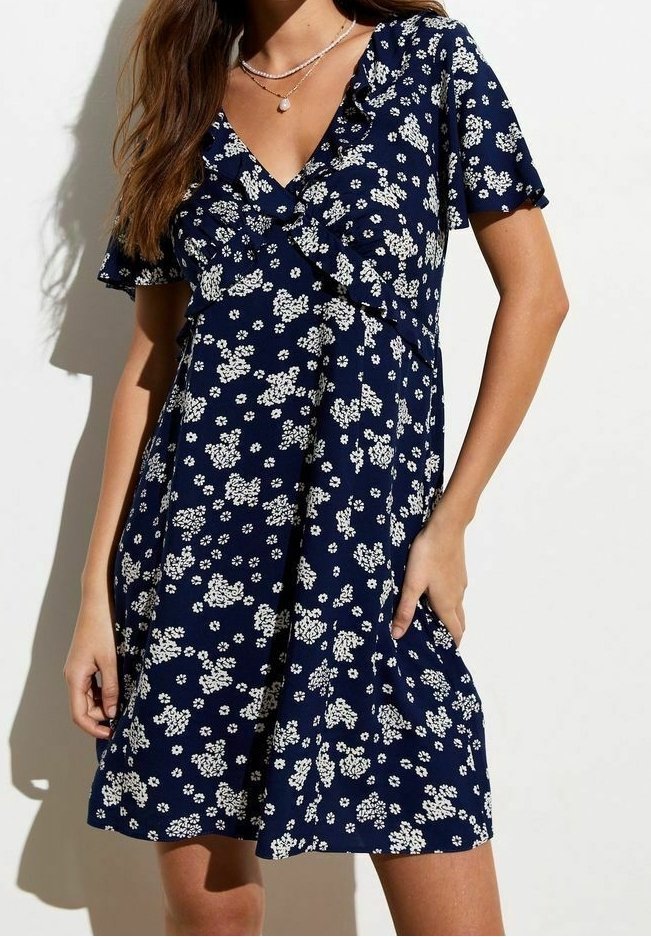 Femme portant une robe bleu marine à motifs floraux, manches courtes et encolure en V, accessoirisée avec des colliers superposés.