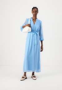 Vaaleansininen pallokuosis maxi-mekko, jossa on sidottava vyötärö, pitkät hihat ja napitusyksityiskohtia. Yhdistetty pieneen valkoiseen käsilaukkuun ja beigeihin kenkiin.