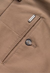 Zbliżenie na jasnobrązową tkaninę o fakturze z pionową kieszenią, ciemnym, okrągłym guzikiem i małą metalową etykietą z napisem "QUINTESSENCE".