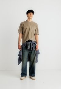 Νέος άντρας που φοράει μπεζ μπλουζάκι, φαρδιά μπλε τζιν, αθλητικά παπούτσια, με ένα καρό πουκάμισο δεμένο γύρω από τη μέση του, μπροστά σε απλό λευκό φόντο.