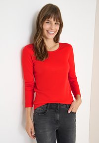 Rotes langärmliges Oberteil mit rundem Ausschnitt und tailliertem Schnitt, kombiniert mit dunkelgrauen Jeans. Glattes Material mit leichtem Glanz.