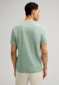 Hellgrünes Baumwoll-T-Shirt mit Rundhalsausschnitt und kurzen Ärmeln, glatte Textur und lockere Passform, getragen von einem Modell, das von der Kamera abgewandt ist.