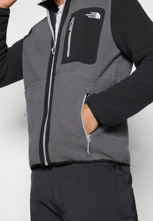 Mann trägt schwarze und graue Fleecejacke mit Reißverschlusstaschen an Brust und Seiten, Hand in Tasche, Logo auf der Brust.