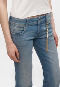 Femme portant un jean en denim bleu et un haut blanc côtelé, avec un accessoire chaîne perlée en or et perles suspendu à la boucle de ceinture du jean.