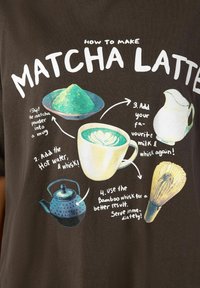 Smeđi majica s raznobojnim grafičkim uputstvima za pravljenje matcha latte. Uključuje ilustracije matcha praha, šalice, kuhala i pjenjače.