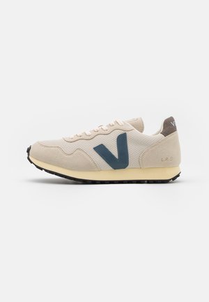 Beiger und cremefarbener Sneaker mit einem großen dunkelblauen V-Logo an der Seite, schwarzer Sohle und weißen Schnürsenkeln auf weißem Hintergrund.