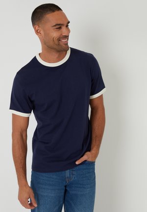 Homme souriant, portant un t-shirt bleu marine avec bordure blanche au col et aux manches, et un jean bleu, debout devant un fond blanc uni.