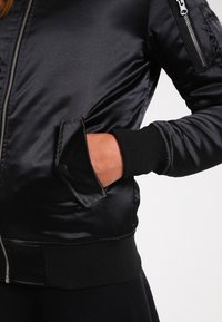 Veste bomber en satin noir avec des poignets et un ourlet côtelés, dotée d'une poche latérale avec rabat et d'une poche zippée sur la manche pour un rangement sécurisé.
