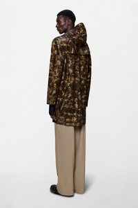 Μακρύ, μακρυμάνικο μπουφάν με κουκούλα σε καφέ και χρυσό σχέδιο tie-dye. Διαθέτει άνετη εφαρμογή, πλαϊνές σκισμές και συνδυάζεται με μπεζ παντελόνι φαρδιάς γραμμής.