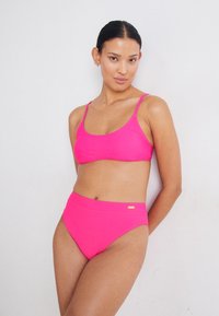 Conjunto de biquíni rosa choque com um top tipo bralette de alças finas e parte de baixo de cintura alta. Textura suave com um pequeno detalhe em metal.