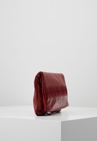 Červená kožená taška s texturovaným vzorem na přední straně, s překlap�ávacím designem a bočním zipem. Hladký povrch a strukturovaný tvar.