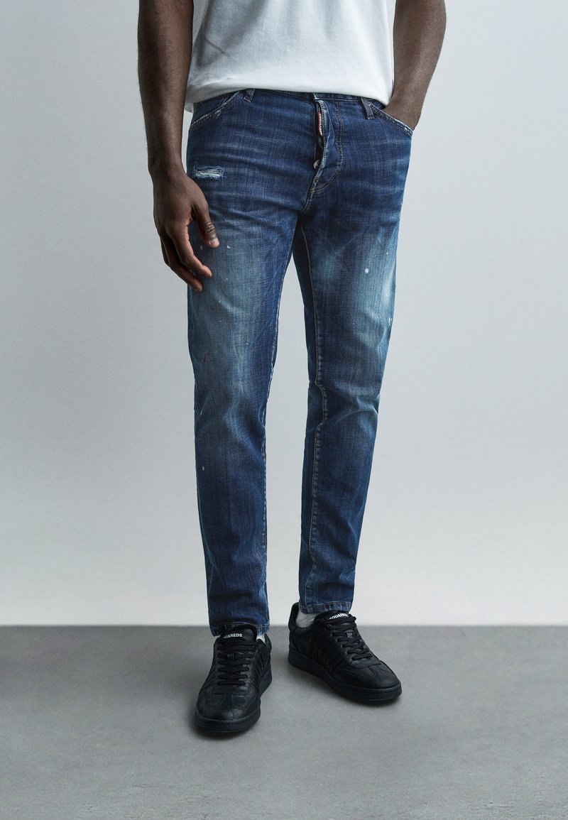Homme portant un jean slim bleu délavé avec des éclaboussures de peinture, des chaussettes blanches et des baskets noires, debout devant un arrière-plan gris.