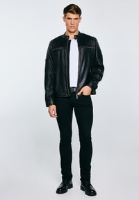 Fiatal férfi áll zsebre tett kézzel, fekete bőrdzsekit, fehér pólót, fekete nadrágot és fekete csizmát viselve világos háttér előtt.