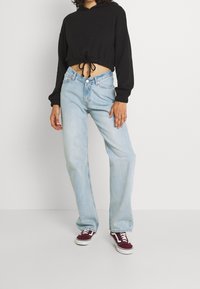 Cappuccio nero corto con cordoncino, jeans dritti azzurri chiari e sneakers basse bordeaux con dettagli bianchi. Tessuto morbido e vestibilità casual.