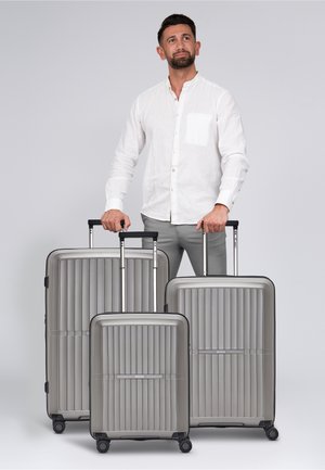 Trois valises métalliques grises avec une texture à rainures verticales et des accents noirs, équipées de poignées rétractables et de roues roulantes.