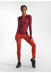 Rotes Langarm-Henley-Shirt mit Knöpfen, kombiniert mit roten Leggings und schwarzen Sportschuhen. Glattes Material mit figurbetontem Design.