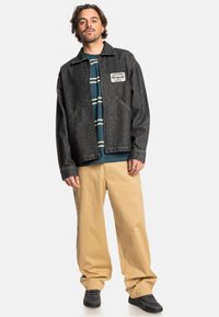 Black denim jacket with a zip closure and two front pockets, worn over a navy striped shirt, paired with beige wide-leg pants and black shoes.