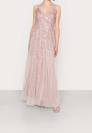 Donna che indossa un abito lungo fino al pavimento color rosa pallido con paillettes, spalline sottili e tacchi argento aperti, in piedi contro uno sfondo chiaro e semplice.