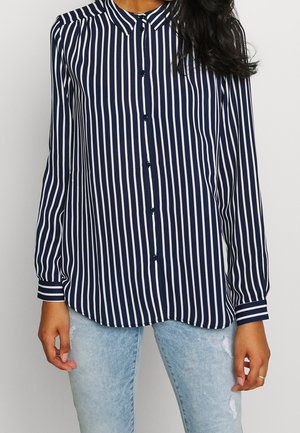 Vrouw draagt een marineblauw-wit verticaal gestreept overhemd met lange mouwen en lichtblauwe versleten spijkerbroek.