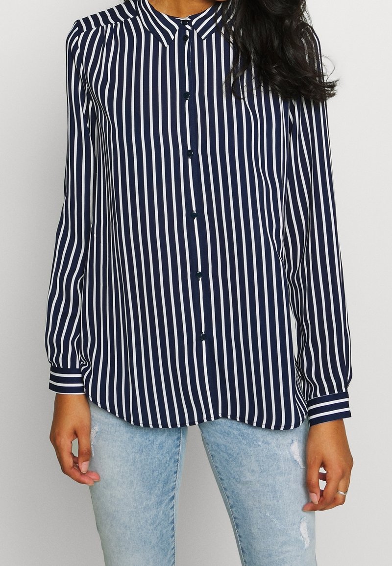 Femme portant une chemise à rayures verticales bleu marine et blanche à manches longues avec un jean bleu clair usé.