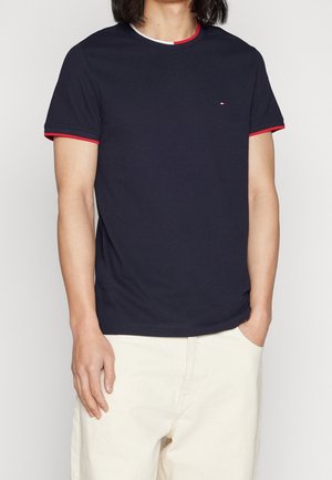 Mand iført en marineblå t-shirt med rød og hvid kant på krave og ærmer, kombineret med off-white bukser, stående med den ene hånd i lommen.