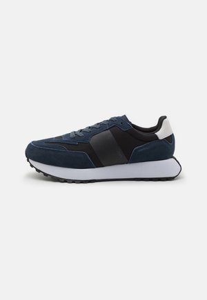 Sneaker in suede blu navy con dettagli neri, sovrapposizioni testurizzate e una suola bianca spessa. Presenta lacci rotondi e una finitura liscia.