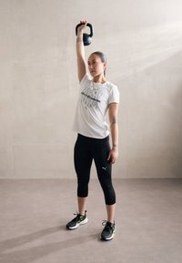 Kettlebell hoiab pea kohal inimene, kes kannab valget T-särki tekstiga, musti kapri leggins'e ja musti tennisetossusid laimi aktsentidega, neutraalse taustaga.