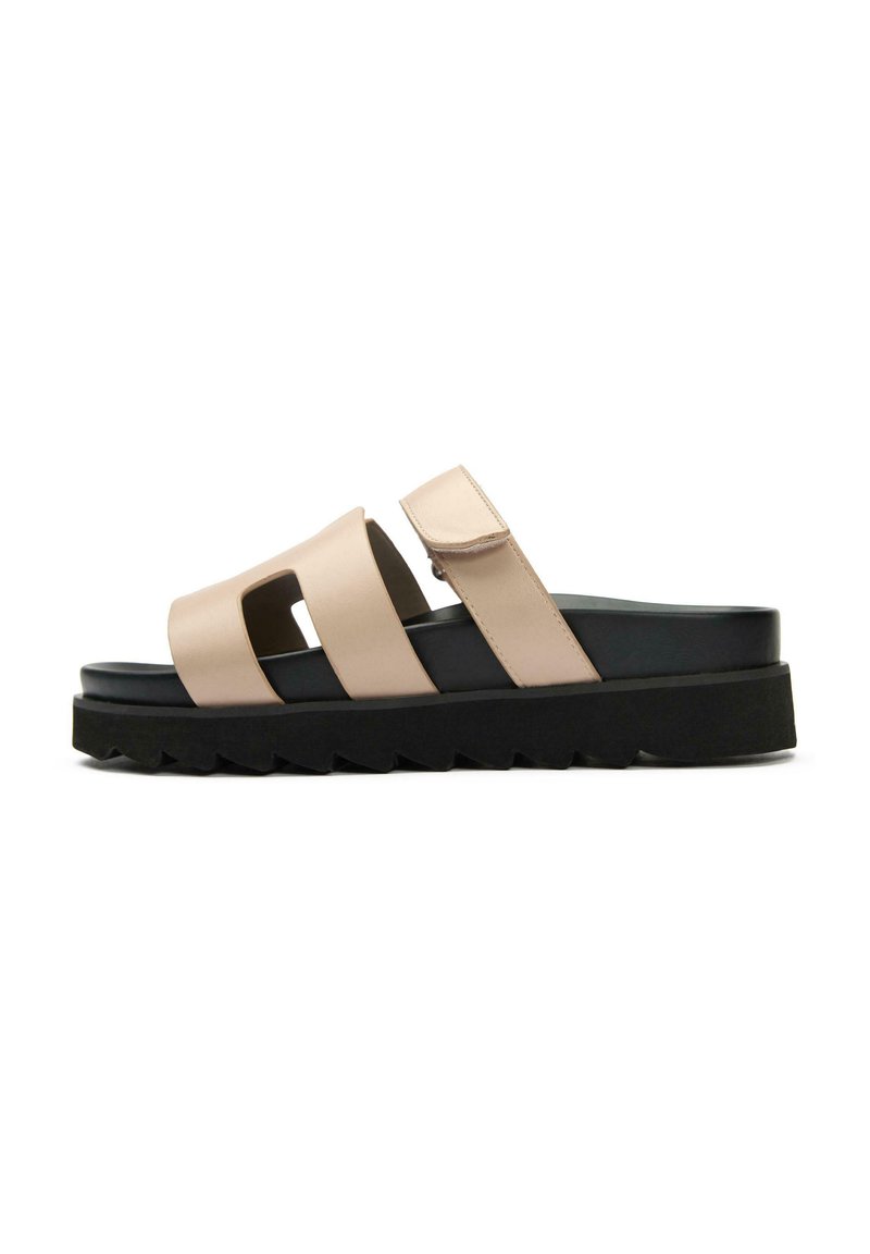 Beige slide sandal with two wide straps and Velcro closure, constructed from smooth material. Black platform sole with a jagged edge.