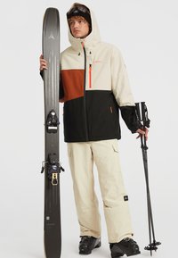 Skiequipment met een beige, zwarte en bruine jas, beige broek en zwarte skibotten, die skies en stokken met paarse accenten vasthoudt.