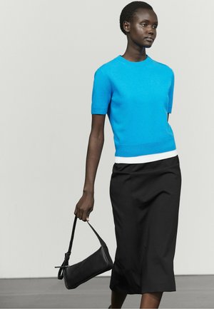 Frau trägt ein leuchtend blaues kurzärmliges Oberteil und einen schwarzen Midirock, hält eine kleine schwarze Handtasche und steht vor einem schlichten hellen Hintergrund.