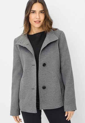 Frau trägt einen grauen Wollmantel mittlerer Länge mit weitem Kragen und schwarzen Knöpfen, kombiniert mit schwarzem Oberteil und Hose, steht vor einem weißen Hintergrund.