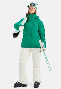 Groene waterdichte skijack met een voorkwini, witte sneeuwbroek, zwarte skischoenen en licht turquoise skilatten in één hand.