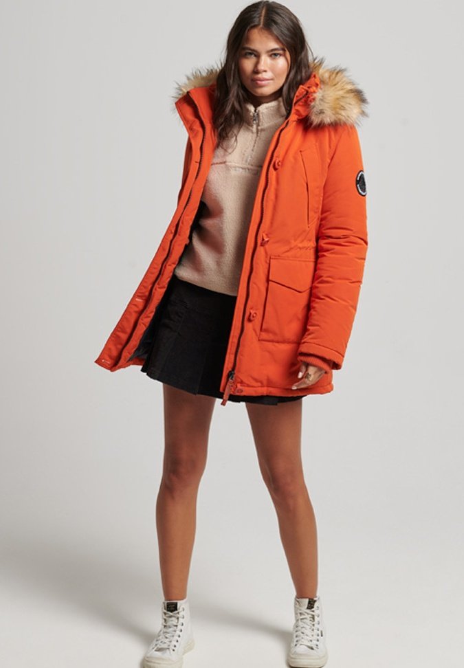Vrouw in een oranje parka met capuchon en bontaccent, beige fleecetop, zwarte rok en witte high-top sneakers, staande tegen een lichte achtergrond.