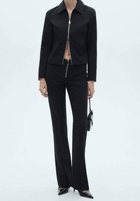 Veste noire courte avec un col pointu et une fermeture éclair argentée, associée à un pantalon noir ajusté et des escarpins à bout pointu, créant un look cintré.