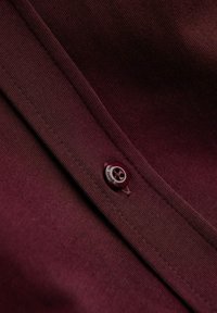 Burgundský hedvábný materiál s texturou, s kulatým tmavým knoflíkem se vzorem lebky a kontrastním prošitím podél švu.
