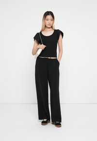 Črn crop top s kratkimi oblazinjenimi rokavi, v kombinaciji s širokimi črnimi hlačami. Model drži majhno kremasto torbico čez ramo. Vidna črna obutev.