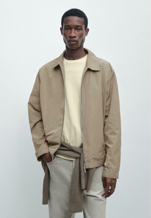 Giovane uomo con giacca beige chiaro sopra maglione crema e maglione marrone a maglia legato in vita, in piedi davanti a uno sfondo bianco uniforme.