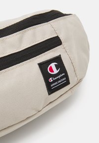 Geantă din material bej, cu un buzunar cu fermoar, având o dungă neagră și un patch cu logo-ul Champion care include textul "HAINE SPORTIVE AUTENTICE."