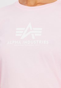 Růžové bavlněné tričko s bílým logem se stylizovaným písmenem "A" a pruhy. Text obsahuje "ALPHA INDUSTRIES" a "KNOXVILLE, TENNESSEE, U.S.A."