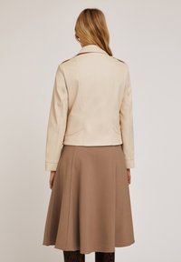 Giacca in similpelle beige dal taglio sartoriale, con colletto a scollo e dettagli delle cuciture in vista, indossata sopra una gonna marrone a ruota.