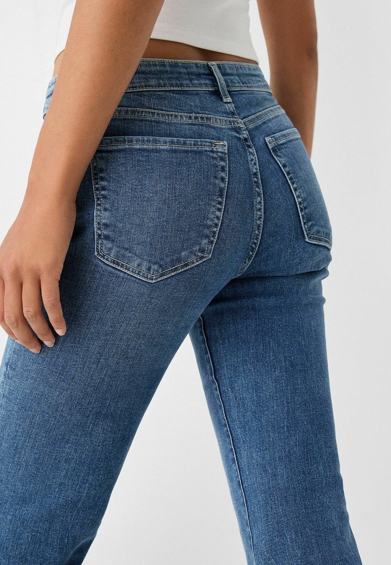 Ingrandimento di una persona che indossa jeans di denim blu e un top corto bianco, con vista sui taschini posteriori e parte della mano posata sulla coscia.