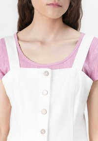 Vestido overall blanco de algodón con cuatro botones metálicos, superpuesto sobre una blusa rosada texturizada con un escote ondulado.