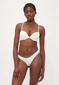 Balts krūšturis ar apakšstīpi un regulējamām lencēm, saskaņots ar baltām bikini biksītēm. Gluda tekstūra un minimālistisks dizains.