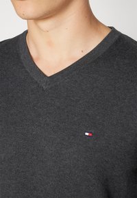 Dunkelgrauer V-Ausschnitt-Pullover aus Strickstoff mit einem kleinen, gestickten Logo in Rot, Weiß und Blau auf der linken Brustseite.