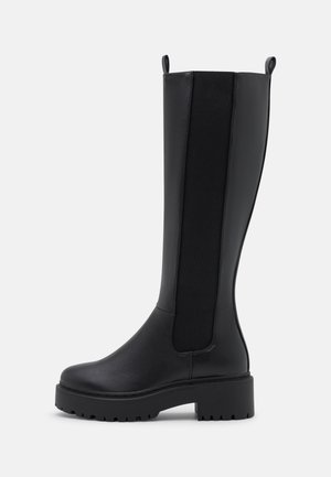 Botte noire montante jusqu’au genou avec semelle épaisse, panneaux latéraux élastiques et languettes sur un fond uni gris clair.